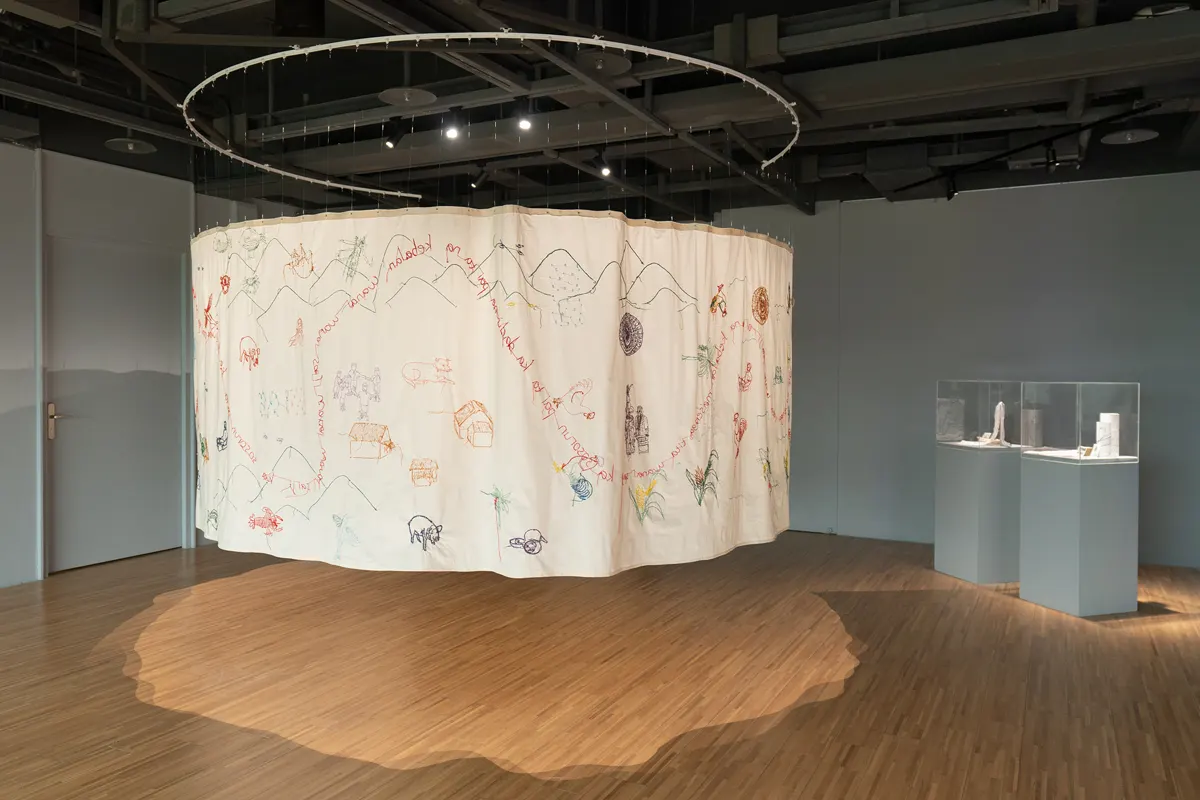 Meita Meilita, „Wanai Saiji: the shape of being“, 2025 m.: kabantis apskritas audinio užuolaidos kūrinys su siuvinėtais kalnais, gyvūnais, namais ir užrašais