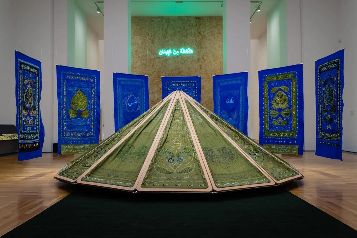 Mounira Al Solh instaliacija: mėlyni siuvinėti audiniai su lapų motyvais, žalia siuvinėta palapinės formos skulptūra ir neoninis užrašas arabų kalba