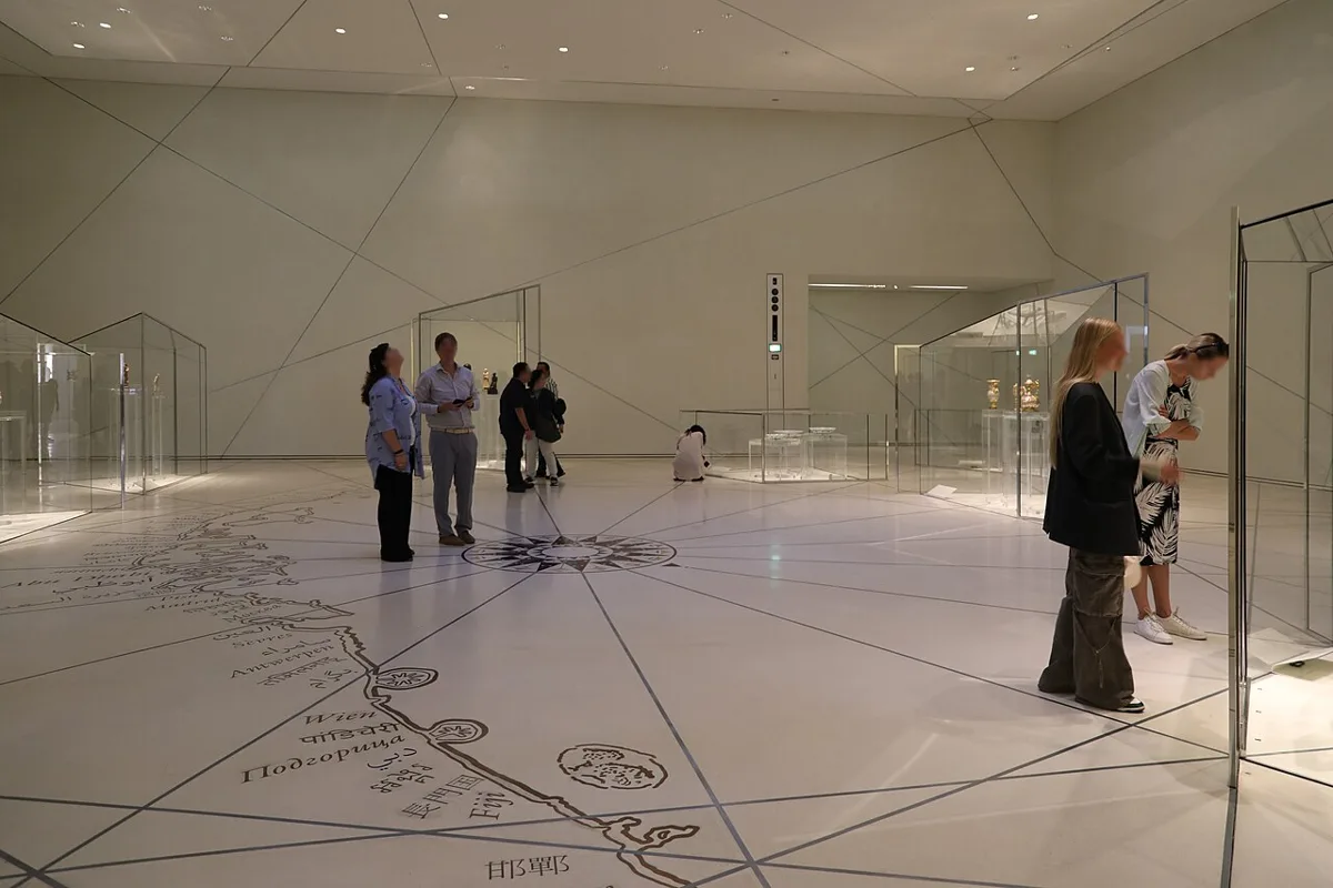Pirmoji Louvre Abu Dhabi nuolatinės ekspozicijos salė: lankytojai prie stiklinių vitrinų, ant grindų pavaizduotas žemėlapis su miestų pavadinimais