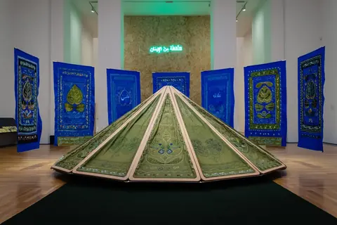 Mounira Al Solh instaliacija: mėlyni siuvinėti audiniai su lapų motyvais, žalia siuvinėta palapinės formos skulptūra ir neoninis užrašas arabų kalba